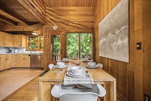 A wooden interior with a dining table set for six, a kitchen with wooden cabinets, and a large window with a view of greenery. - Holzhutte - Mid-century alpine retreat - Hot tub (Government Camp)