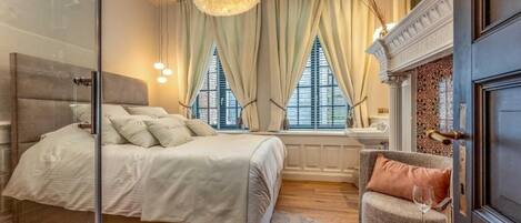 Ein Schlafzimmer mit einem Bett, einem Sessel, einem Kamin und einem Fenster mit Vorhängen.