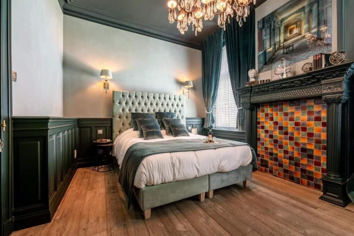 Une chambre à coucher avec une tête de lit capitonnée, un grand lit, un lustre, une cheminée ornée de carreaux colorés et un tableau au-dessus de la cheminée.
