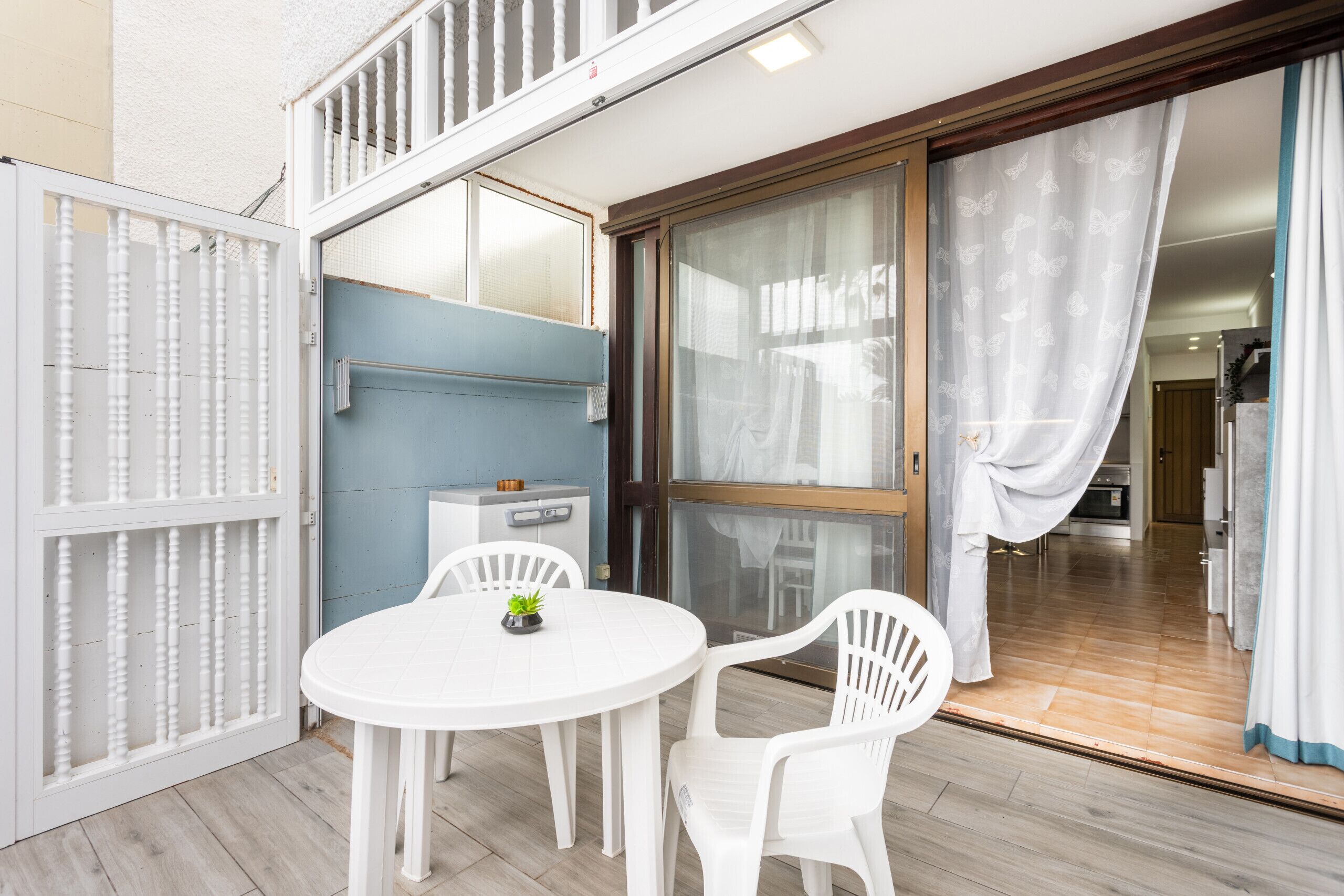 Une pièce avec une table ronde, deux chaises, une petite plante, un mur bleu, une porte-fenêtre coulissante et un coin cuisine visible à travers la porte.