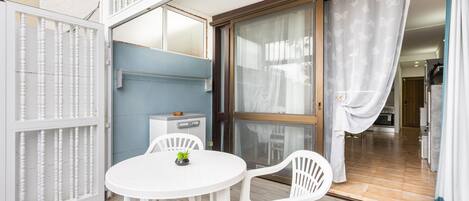 Une pièce avec une table ronde, deux chaises, une petite plante, un mur bleu, une porte-fenêtre coulissante et un coin cuisine visible à travers la porte.