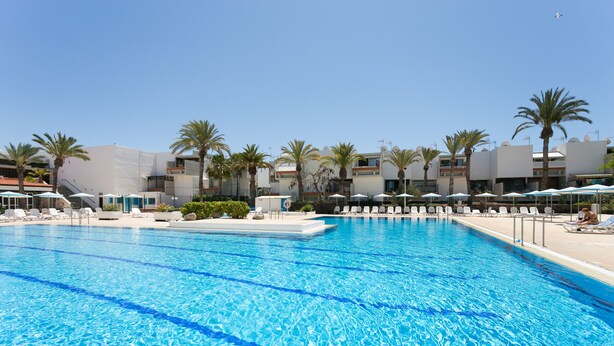 Una piscina con acqua cristallina e blu, circondata da sdraio e ombrelloni, con palme e edifici moderni sullo sfondo.