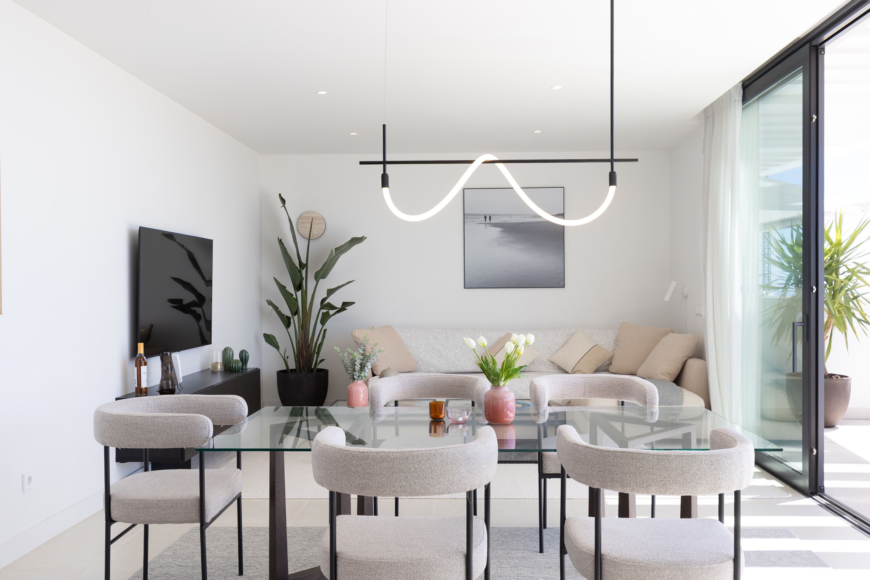 A modern living room with a glass dining table, grey upholstered chairs, a sofa with beige cushions, a large potted plant, and a wall-mounted TV.