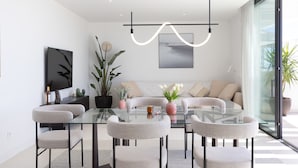 Un soggiorno moderno con un tavolo da pranzo in vetro, poltrone rivestite di grigio, un divano con cuscini color beige, una grande pianta in vaso e una televisione a parete.