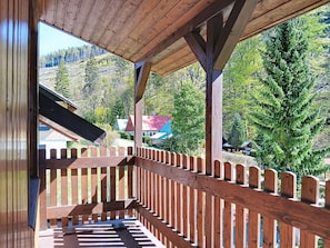 A wooden balcony with a view of a forested mountain area and a red-roofed building.