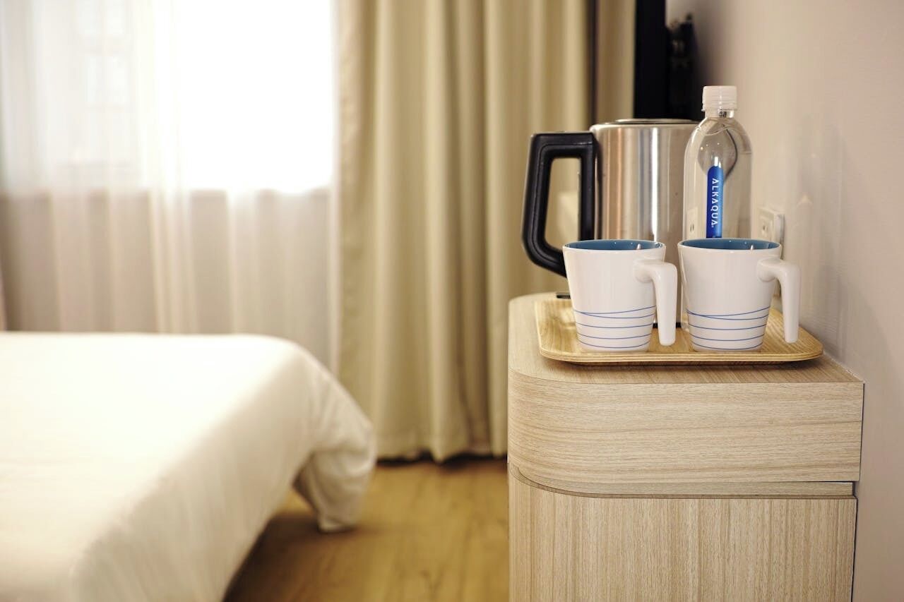 A hotel room with a bed, a bedside table with a kettle, a water bottle, and two mugs.