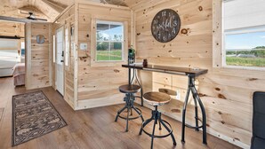 A wooden interior with a kitchen area, including a wooden table and stools, a clock on the wall, and a view through the window. - Cozy stay at Baling Out Farms (Southside)