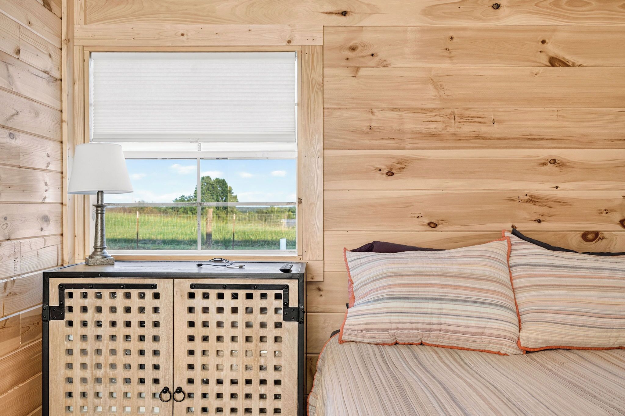 A wooden cabin interior with a bed, a window with a view of a green field, and a radiator.
