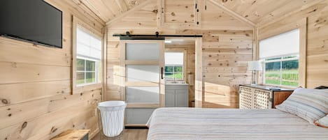 A wooden cabin interior with a bed, a bench, a TV, a window, and a door leading outside.