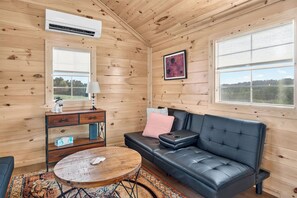 Interior acogedor de una cabaña con un sofá de cuero, una mesita redonda, un aparador y una ventana con vistas.