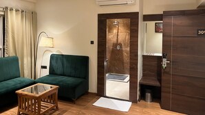 A hotel room with a green sofa, a wooden coffee table, a glass shower enclosure, an air conditioning unit, and a door with a number plate.