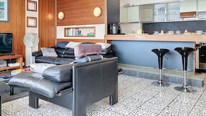 Un salon moderne comprenant un canapé, une table basse, un téléviseur et un ventilateur. Il y a également un coin cuisine avec un comptoir et des tabourets.