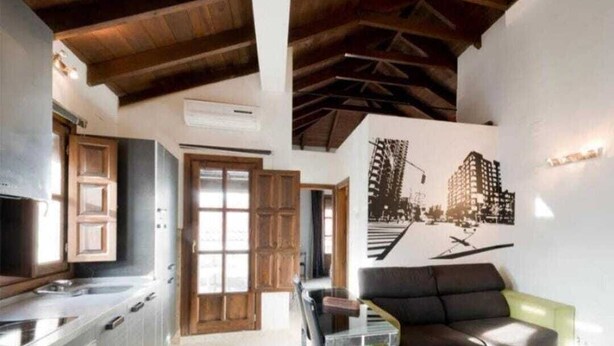 A modern kitchen with wooden cabinets, a dining area with a sofa and a coffee table, and a wall with a cityscape mural. - Spectacular penthouse with private terrace well communicated in the center of Granada (Granad)