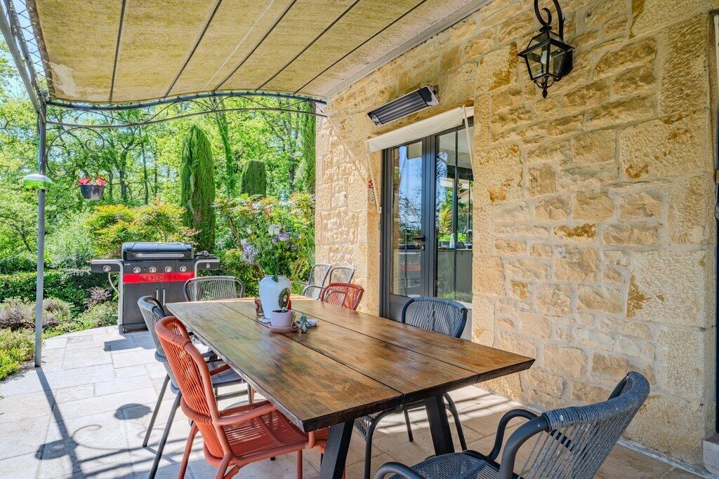 Een terras buiten met een stenen muur, een houten tafel, rieten stoelen, een barbecue en hangplanten.