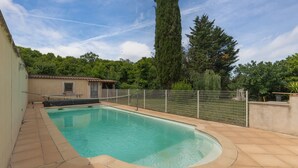 A swimming pool with a tiled deck, a fenced area, and a tall tree in the background.