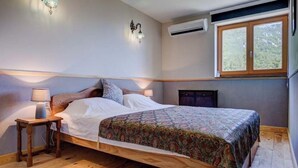Une chambre avec un lit en bois, une table de chevet avec une lampe, un climatiseur fixé au mur et une fenêtre donnant sur des arbres.