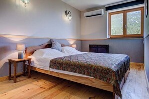 A bedroom with a wooden bed, a nightstand with a lamp, a wall-mounted air conditioner, and a window with a view of trees. - Peaceful luxury with fabulous views. (Budva)