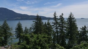 A coastal landscape with dense coniferous trees, a calm body of water, and distant mountains under a clear sky. - Exclusive 2-bdrm ocean & mountain view suite (Bowen Island)