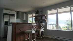 A kitchen with a wooden island, two bar stools, a refrigerator, and large windows offering a view. - Exclusive 2-bdrm ocean & mountain view suite (Bowen Island)