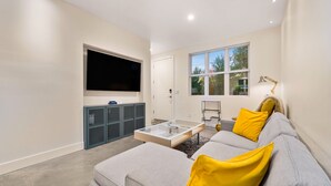 Ein modernes Wohnzimmer mit Flachbildfernseher, grauem Sofa mit gelben Kissen, einem Couchtisch und einem Fenster mit Blick auf Bäume.