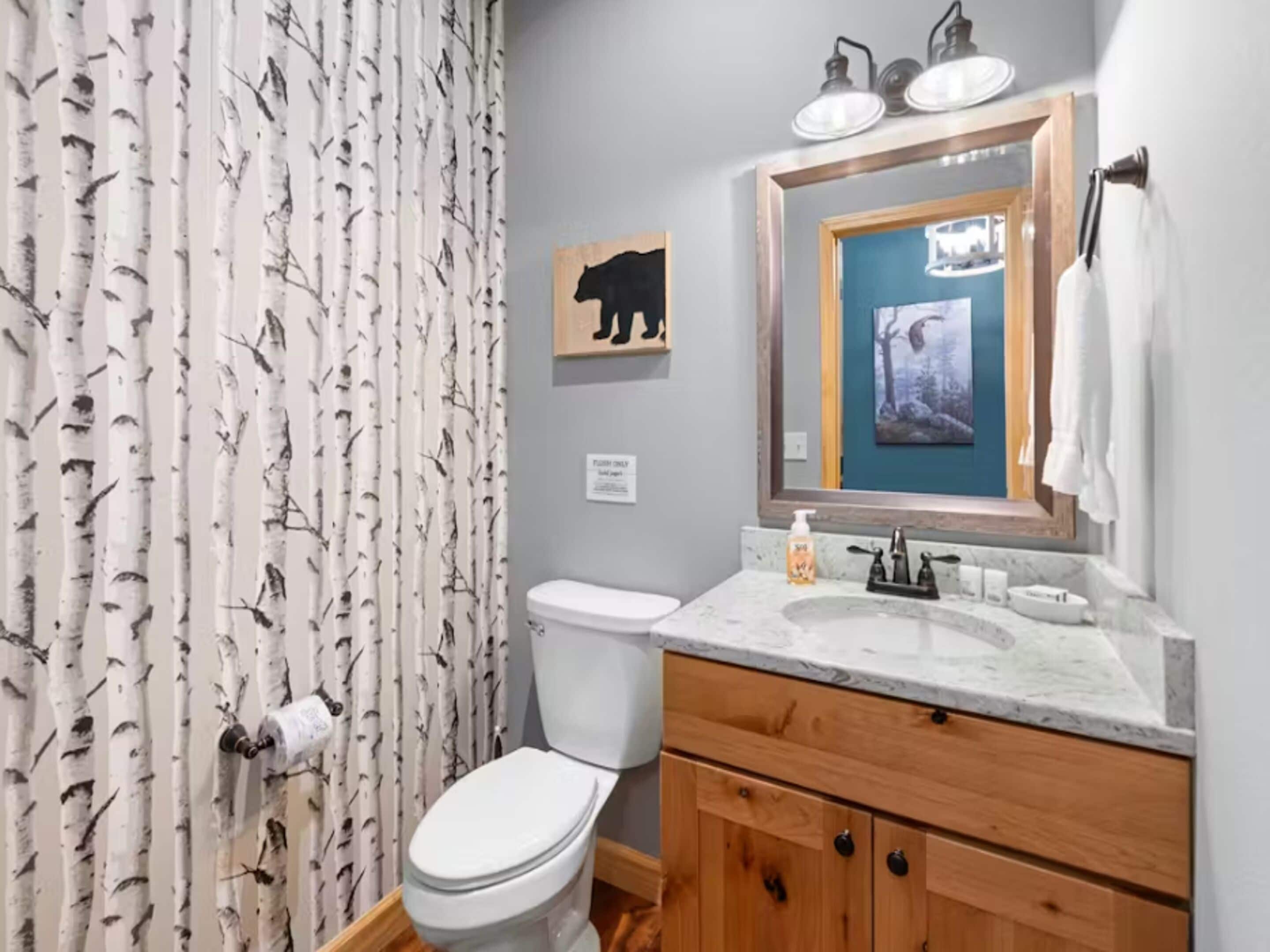 A bathroom with a toilet, a sink with a mirror, a shower curtain with a tree pattern, and a wall-mounted sign.