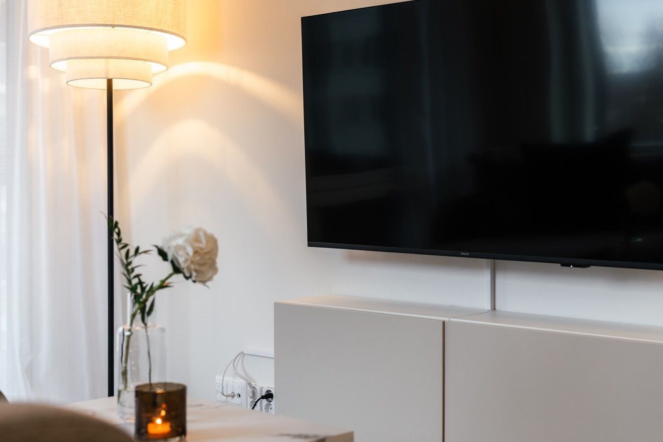 Ein modernes Wohnzimmer mit einem an der Wand montierten Flachbildfernseher, einem weißen Schrank darunter, einer Stehlampe und einer Vase mit Blumen auf einem Tisch.