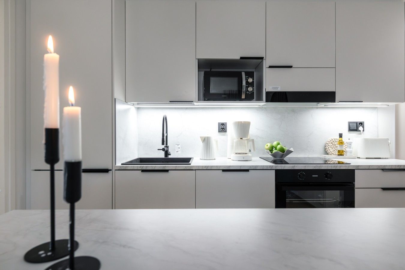 A modern kitchen with a built-in microwave, a sink, and a stove. There are candles on a stand, a coffee maker, and a bowl of green apples on the counter.