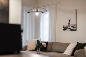 A living room with a sofa, a decorative light fixture, a framed picture on the wall, and a window with sheer curtains. - Snownest Premium City Centre Apartments 2 (Rovaniemi)