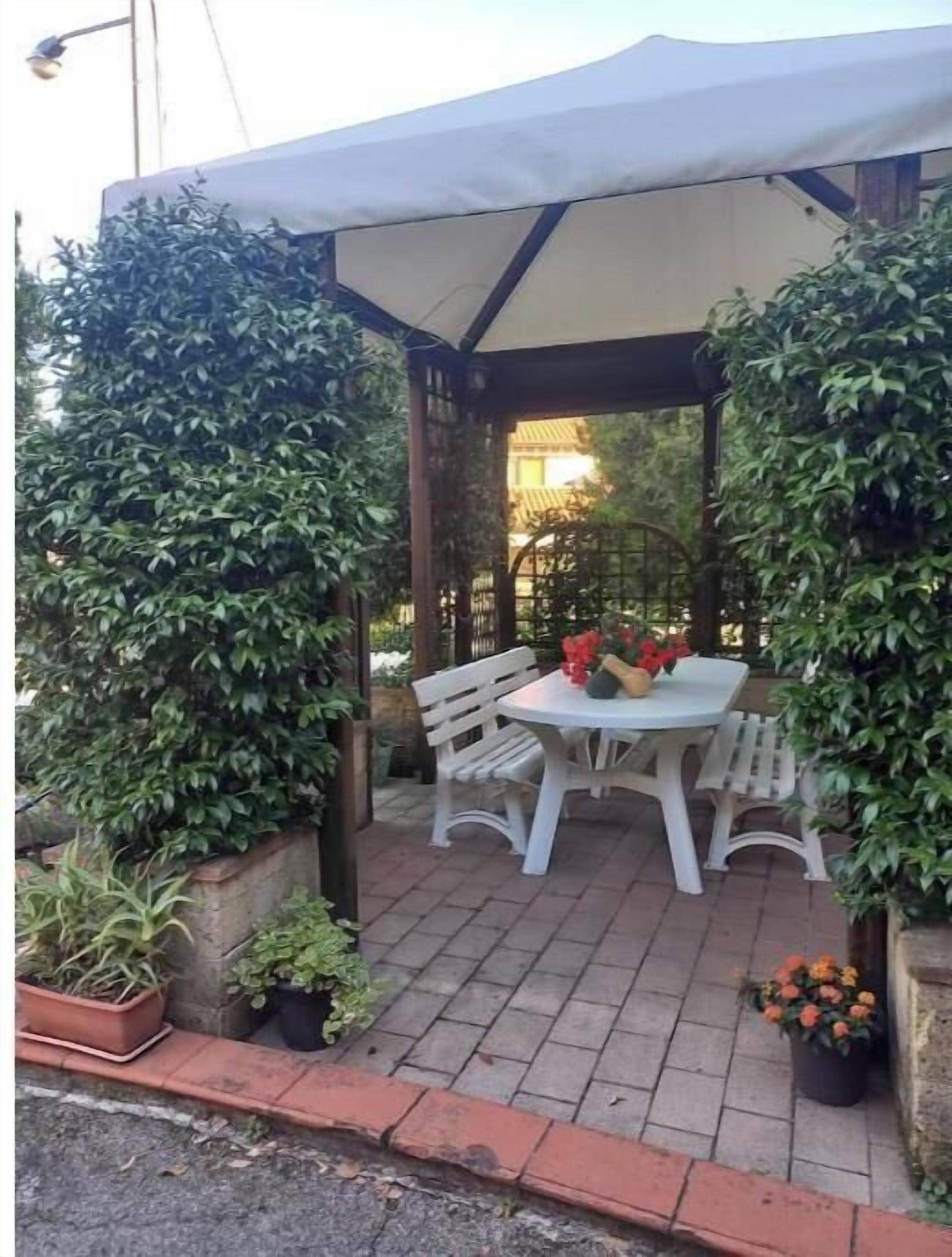 A covered patio area with a table and chairs, surrounded by potted plants and a pergola structure.