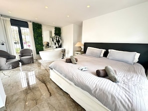 A modern bedroom with a large bed, a sitting area with a chair, a bathtub, and a view of greenery outside. - 3305 Carib Playa (Marbella)