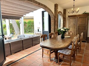 A dining area with a wooden table, chairs, and a bench. There is a chandelier above the table and a view of the outdoors through large windows. - 3305 Carib Playa (Marbella)