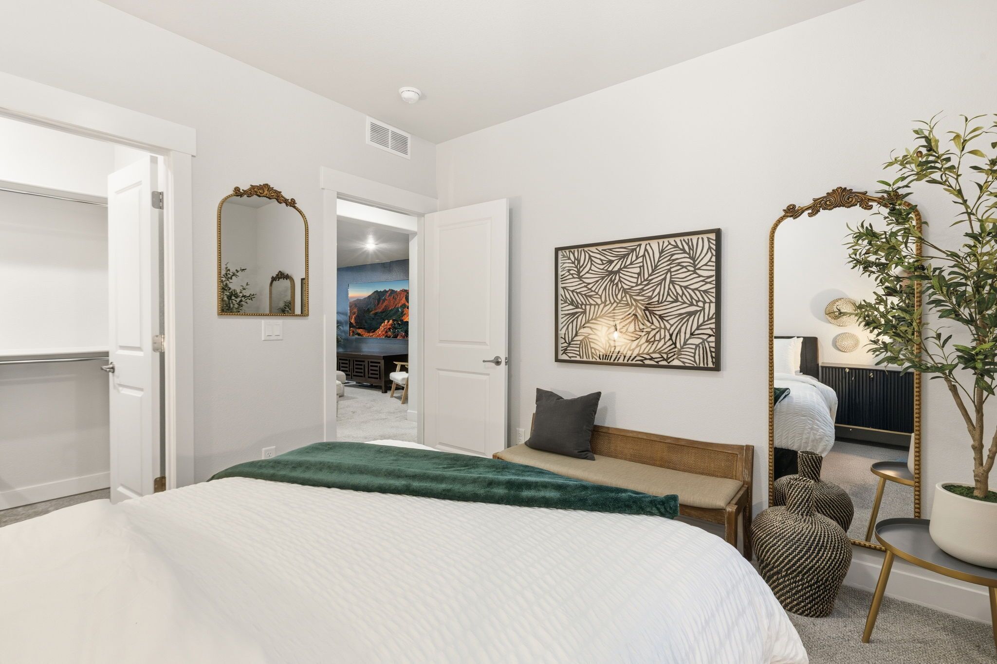 A bedroom with a bed, a mirror, a plant, and a view of another room.