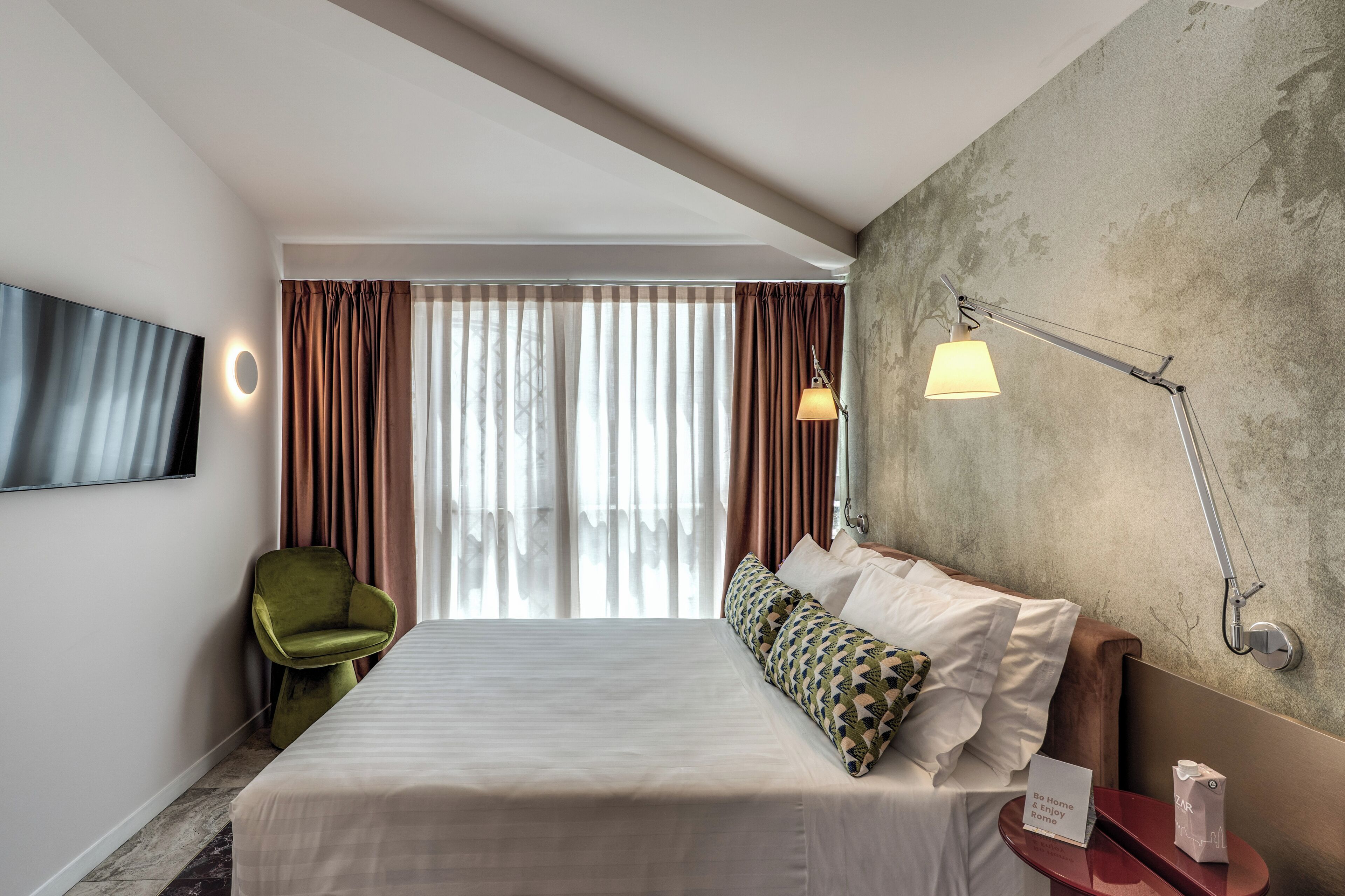 Una camera d'albergo con un letto, una poltrona verde, un comodino, una lampada e un'ampia finestra con tende.