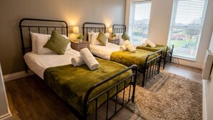 Ein Zimmer mit drei Betten, jeweils mit Metallgestell und grüner Bettwäsche. Nachttische mit Lampen sind vorhanden, und das Zimmer verfügt über große Fenster mit Jalousien.