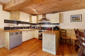 A kitchen with wooden cabinets, a built-in dishwasher, a stove, and a dining area with a wooden table and chairs. - Puddle-duck Cottage – La Mare Chappey, children welcome, heated pool (Néhou)