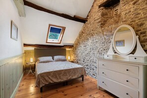 A bedroom with a stone wall, a bed with a patterned bedspread, a bedside table with a lamp, and a dresser with a mirror. - Gîte Grange – La Mare Chappey, child-friendly, heated pool (Néhou)