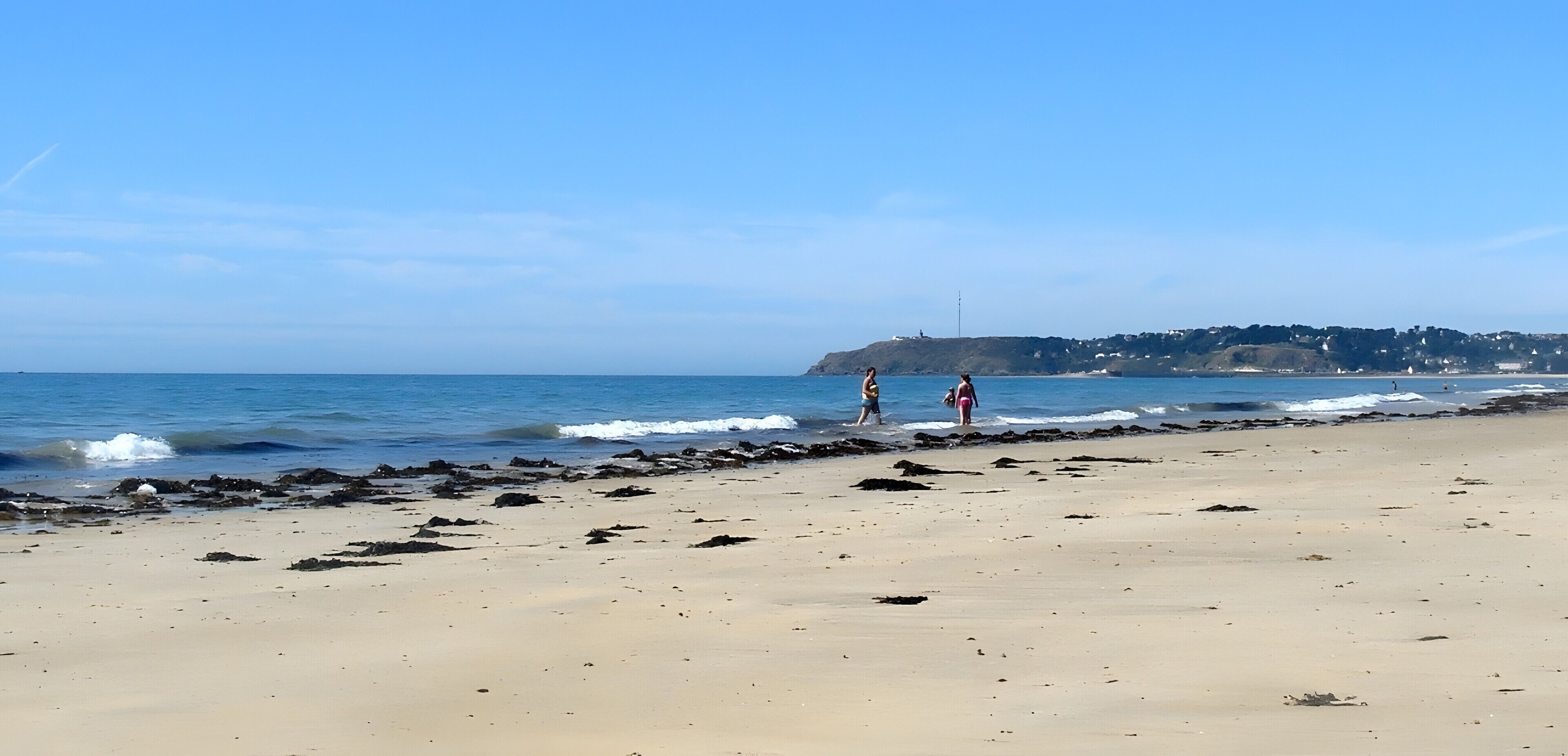 Une plage bordée d’une côte rocheuse, une mer calme et, au loin, une colline parsemée de bâtiments.