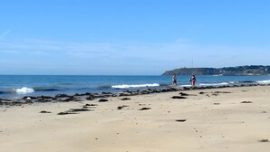 A beach with a rocky shoreline, calm sea, and a distant hill with buildings. - Gîte Grange – La Mare Chappey, child-friendly, heated pool (Néhou)