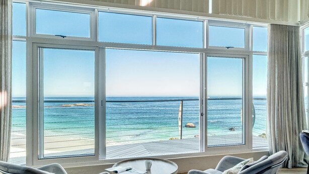A room with a large window overlooking a beach, a small round table, and two chairs. - Camps Bay 6bed w Ocean Views and Pool Villa Sevara (Cape Town)