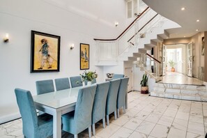 Een eetruimte met een glazen tafel en blauwe fauteuils, een trap en een gang met een potplant.