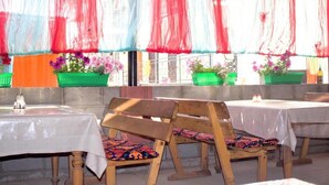 Een buitenterras met houten stoelen en tafels, plantenbakken en uitzicht door een raam met kleurrijke stof.