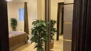 Quarto de hotel com uma cama, uma planta em vaso, um espelho e uma porta que dá para outro cômodo.