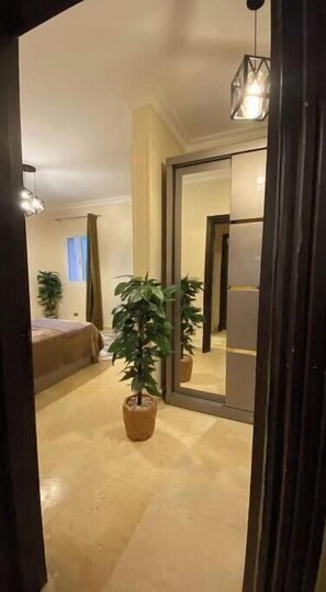 A hotel room with a bed, a potted plant, a mirror, and a door leading to another room. - Luxurious Pool View Apartment – Elegant 2BR in Dreamland Compound (Giza Governorate)