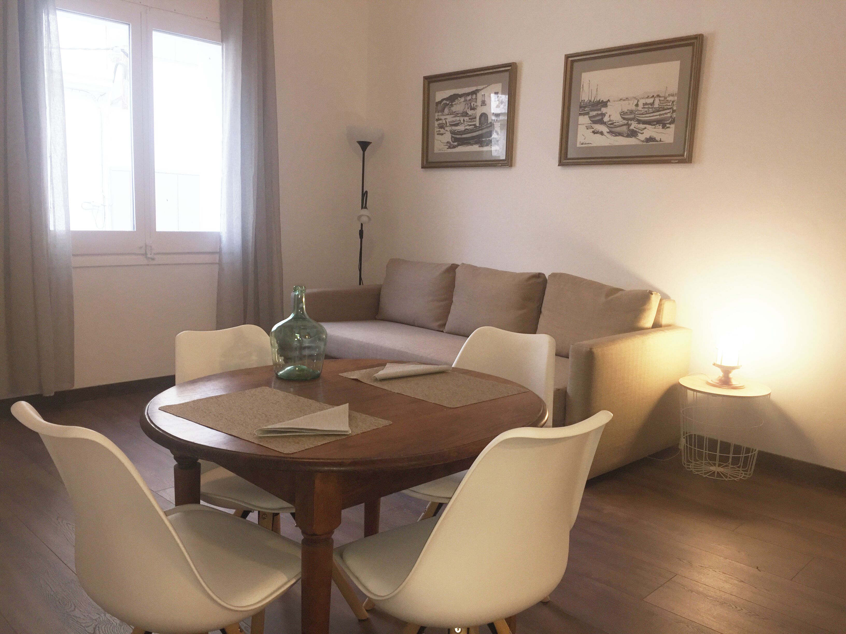 Un comedor con una mesa redonda, cuatro sillas, un sofá, dos cuadros en la pared y una lámpara sobre una mesita auxiliar.