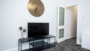 A modern living room with a flat-screen TV on a black glass console, a round wall clock, and a door leading to another room. - St Andrews By Guestz (Lytham Saint Annes)