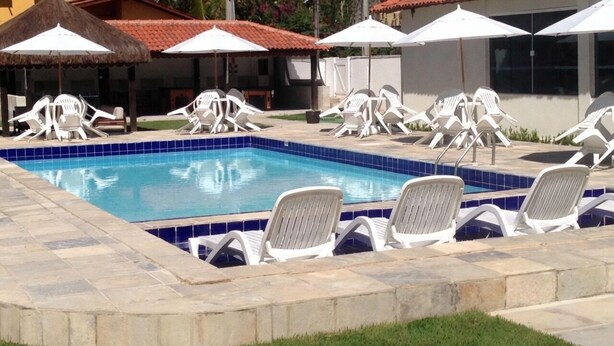 A swimming pool with lounge chairs and umbrellas, surrounded by a stone deck and grass. - Pousada Suites de Porto (Ipojuca)