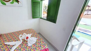 A hotel room with a bed, a window with green shutters, a view of a swimming pool, and a painting on the wall. - Pousada Suites de Porto (Ipojuca)
