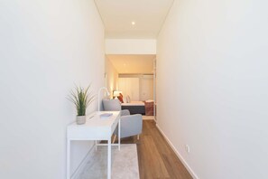 Un pasillo estrecho con un escritorio blanco, una silla gris, una planta en maceta y una cama al fondo.