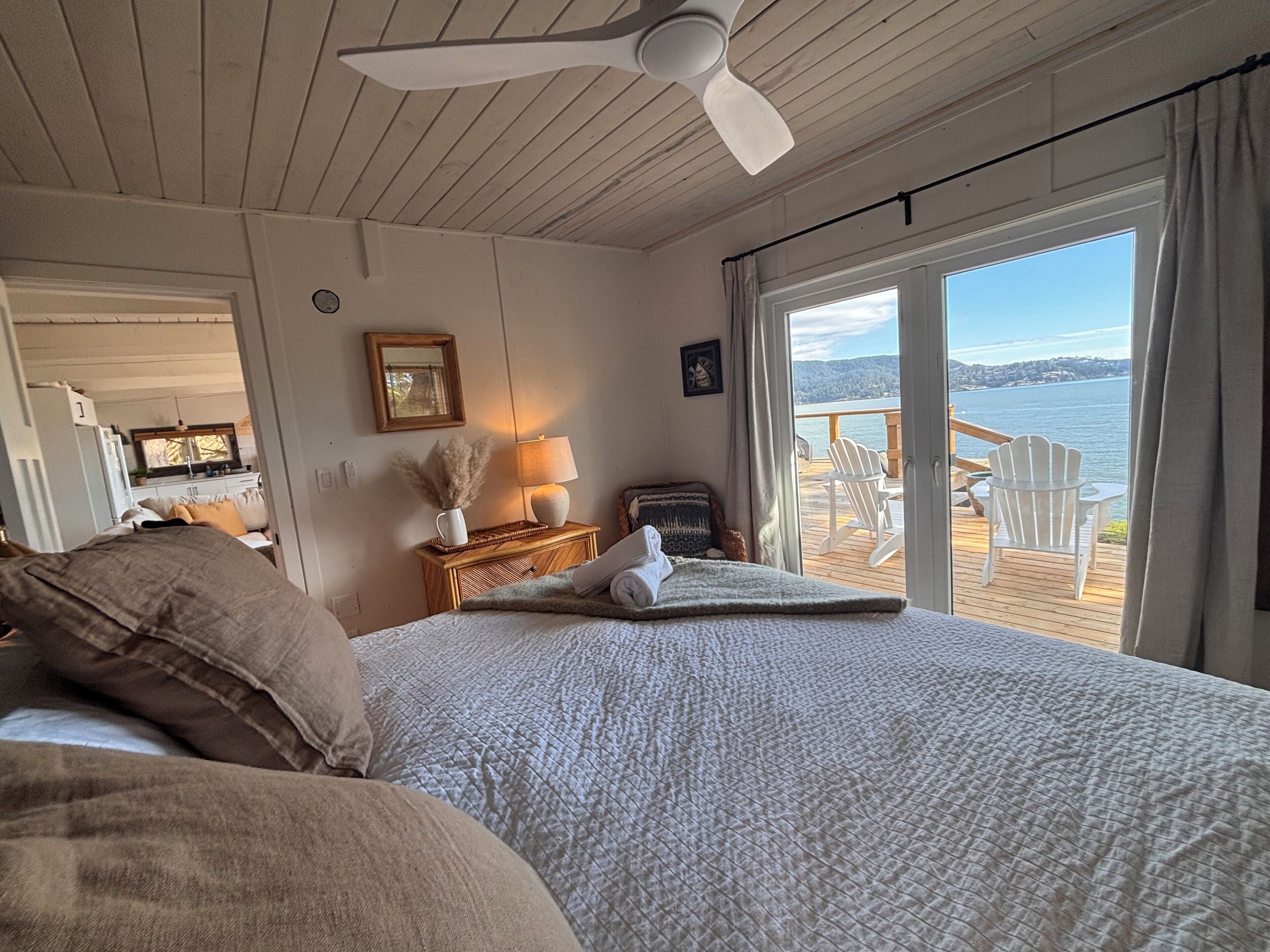 A bedroom with a bed, a nightstand, a lamp, a window with a view of the water, and a ceiling fan.