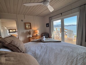 Ein Schlafzimmer mit einem Bett, einem Nachttisch, einer Lampe, einem Fenster mit Blick auf das Wasser und einem Deckenventilator.